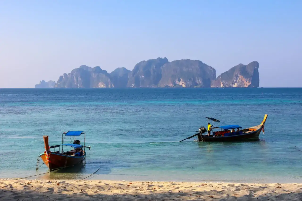 Dwa tradycyjne tajskie łodzie dryfują na turkusowej wodzie. W tle widać skaliste wyspy. Idealne miejsce na wakacje w Tajlandii, Phuket.